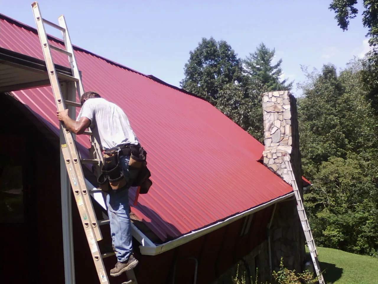 Roofer on a ladder beside a red metal roof and stone chimney — Soffit & Fascia Repair by Roofing Brothers in Johnston